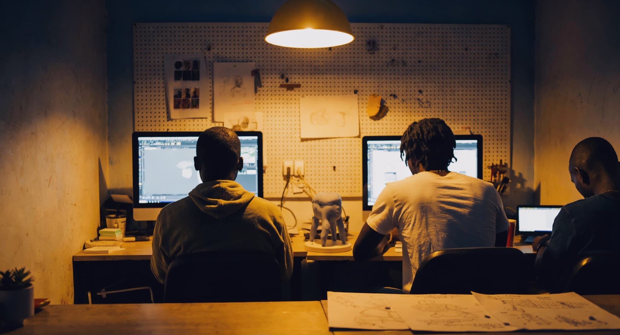 Three people at desks in a small studio under a warm pendant lamp, a pegboard of sketches behind them and a 3D-printed model on the desk